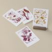 Four artistic floral prints on paper: two red and two gold. Each features detailed illustrations of various flowers, placed on a beige surface. One print is upright in a wooden holder.