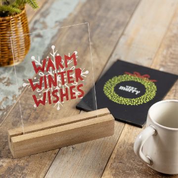 Show details for Reusable Acrylic Bundle A decorative sign with "Warm Winter Wishes" in red and white rests on a wooden holder. Nearby is a black card with a holiday wreath and a white mug on a wooden table.