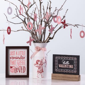 Show details for XO Valentine's Day A decorative setup with a white vase holding branches adorned with pink and red X and O ornaments. Framed signs read "Just a little reminder that you are loved" and "Hello Valentine." The background is white.