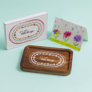 Show details for It's The Little Things A decorative setup featuring a wooden tray and a wall sign, both inscribed with "It's the little things." The tray is brown with intricate white patterns. A colorful, hand-drawn card with flowers is also displayed. Pale green background.