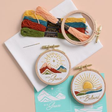 Show details for His Grace Embroidery kit with colorful threads, a needle, two hoops with mountain and sun designs labeled "His Grace" and "His Beloved," fabric, and a stencil. Set on a beige surface.
