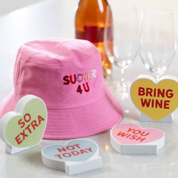 Show details for Sassy Candy Hearts A pink bucket hat with "SUCKER 4 U" embroidered on it is surrounded by heart-shaped signs with phrases like "SO EXTRA," "BRING WINE," "NOT TODAY," and "YOU WISH." Two wine glasses and a bottle are in the background.