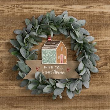Show details for I Am Home A decorative wreath with green leaves surrounds a small house illustration. Below the house, a banner reads "With you, I am home," accompanied by two red hearts. The background is a wooden surface.