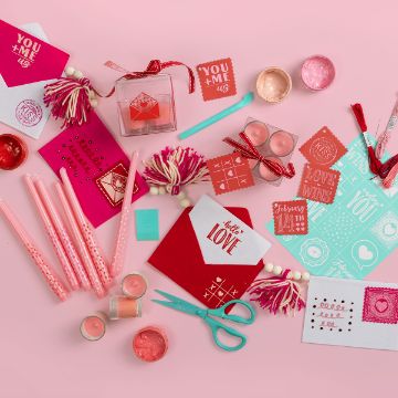 Show details for Sealed With A Kiss A creative Valentine's Day crafting setup with red and pink-themed materials, including cards, scissors, ribbons, stickers, pom-poms, and candles arranged on a pink background.