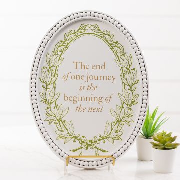 Show details for The Journey An oval sign with a beaded border displays the text "The end of one journey is the beginning of the next," surrounded by a green leaf wreath. The sign is placed on a stand next to two small potted plants.