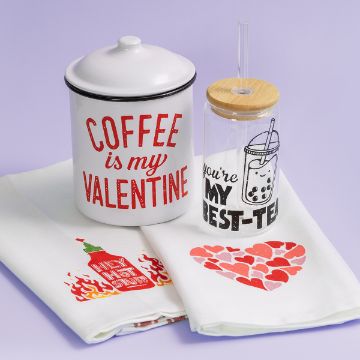 Show details for Hey Hot Stuff A white container with "Coffee is my Valentine" text, a clear tumbler with "You're my best-tea" and bubble tea graphic, and two towels—one with a heart design and the other with a hot sauce bottle. The background is light purple.