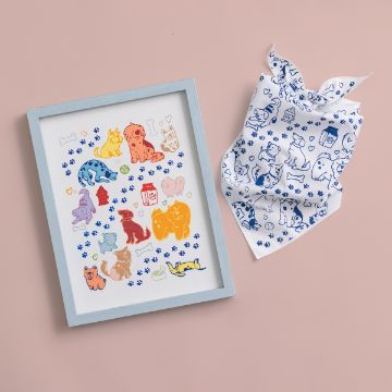 Show details for Triangle Bandana Framed artwork and a patterned cloth on a pink surface. The artwork features colorful illustrations of various dogs in different poses. The cloth has a white background with blue outlines of dogs and paw prints.