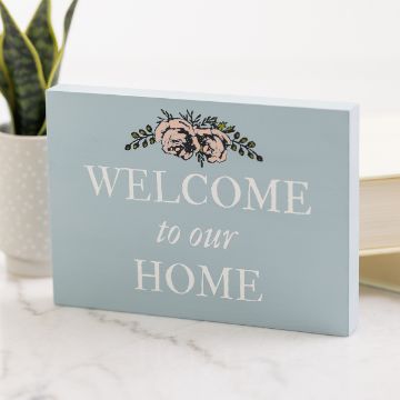 Show details for Chubby Rectangle (5" x 7") A light blue wooden plaque with "Welcome to our Home" written in white text. Above the text is a floral design with pink and green flowers. The plaque is placed on a marble surface near a small plant and a book.
