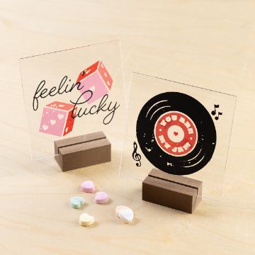 Show details for Acrylic Square (5" x 5", 3 mm) Two small acrylic signs on wooden bases. One features pink dice with "feelin lucky" text. The other shows a vinyl record with "you are my jam" text. Colorful heart-shaped candies lie in front, all on a light wood surface.