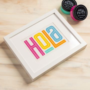 Show details for Hola A framed print displaying the word "HOLA" in bold, colorful letters sits on a light wooden surface. Two small containers of chalk paste, one turquoise and one pink, are nearby.