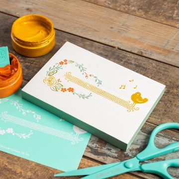 Show details for Songbird A decorated wooden plaque with floral patterns, musical notes, and a bird is on a wooden surface. Nearby are open jars of yellow and orange paint, a piece of teal paper with designs, and a pair of teal scissors.