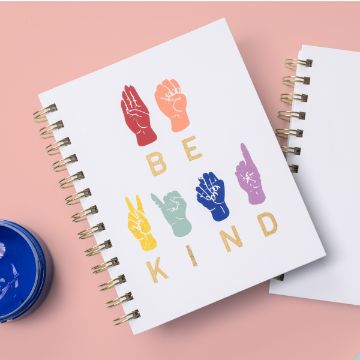 Show details for ASL Alphabet A spiral notebook on a pink surface features colorful hand signs spelling "BE KIND" using American Sign Language. A small jar of blue paint is partially visible beside the notebook.