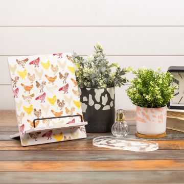 Show details for Chickens Pattern A decorative book with a colorful chicken pattern is displayed on a wooden table, accompanied by two small potted plants, a clear glass jar, and a clock partially visible in the corner. The setup is against a white shiplap wall.
