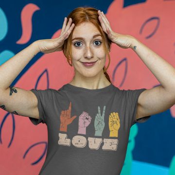 Show details for ASL Love A woman with red hair smiles, holding her hands at her temples. She's wearing a gray T-shirt with "LOVE" spelled in different colored hand signs. The background features a colorful abstract mural.