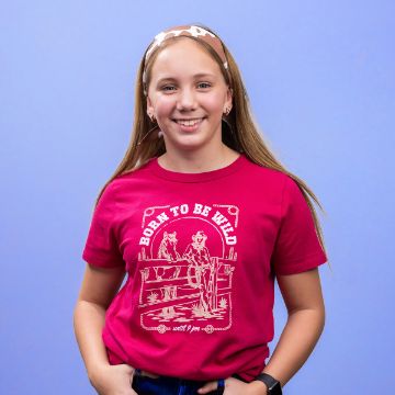 Show details for Born To Be Wild A girl with long hair smiles while wearing a pink T-shirt with a cowboy design and the phrase "Born to be Wild." She stands against a blue background, with her hands in her pockets.