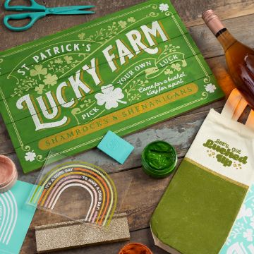 Show details for Lucked Up A flat lay of St. Patrick's themed items: a green "Lucky Farm" sign, rainbow stickers, a "Let's get Lucked Up" tote bag, a green candle, crafting tools, a wine bottle, paint, and a greeting card, all on a wooden surface.