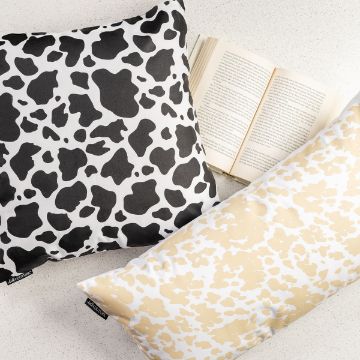 Show details for Cow Print Two pillows with cow print designs, one black and white and the other beige and white, are placed on a light surface next to an open book. The patterns are bold and eye-catching.