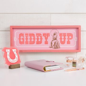 Show details for Giddy Up A pink framed sign displays the text "GIDDY UP" with a silhouette of a cowboy. It's placed on a white table with a pink notebook, a pen, a clear tape dispenser, and a small horseshoe art piece. The background is white wooden panels.