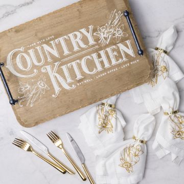 Show details for Made With Love A wooden tray with "Country Kitchen" inscribed, accompanied by floral designs. Below are five gold-accented white napkins with "Love" and "Laugh" bands, alongside gold and silver cutlery on a marble surface.