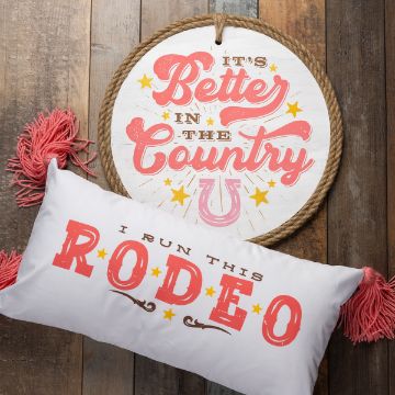 Show details for In The Country A round wooden sign with "It's Better in the Country" in pink and gold lettering hangs on a rustic wooden wall. Below it, a white pillow with red tassels reads "I Run This Rodeo" in bold red letters with stars.