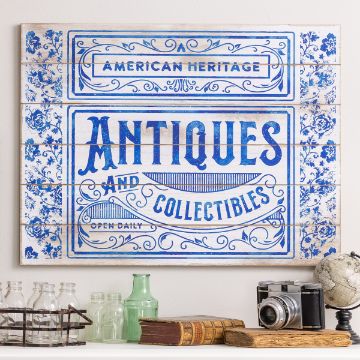 Show details for Antiques A decorative sign with blue lettering reads "American Heritage Antiques and Collectibles, Open Daily" on a whitewashed wooden background with floral motifs. Below are glass bottles, books, a camera, and a globe displayed on a shelf.