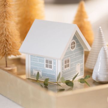 Show details for Cozy Cottage A small decorative model of a blue and white house with a triangular roof is placed on a tray. Surrounding it are miniature tan and white cone-shaped trees, creating a cozy, festive scene. Greenery adds a touch of natural decor.