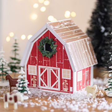 Show details for Country Barn A festive red and white barn model decorated with a green wreath is surrounded by small pine trees, artificial snow, and toy animals. Soft, blurred lights twinkle in the background, creating a cozy holiday atmosphere.