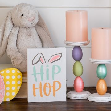 Show details for Hip Hop A plush rabbit sits beside a small sign that reads "Hip Hop" with bunny ears illustration. Nearby, two stands hold stacked pastel-colored eggs topped with pink candles. A wooden egg with polka dots rests on the left.