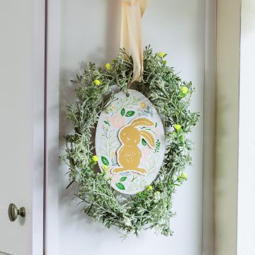 Show details for Storybook Bunny A decorative wreath hangs on a door, featuring green foliage with small yellow flowers. In the center, there's an oval placard with a cute yellow rabbit illustration surrounded by floral designs. A beige ribbon holds the wreath in place.