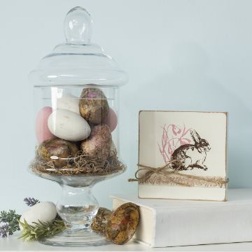 Show details for Best Easter Wishes A glass jar filled with decorated Easter eggs on a bed of straw sits beside a square plaque featuring a rabbit design. The setup is on a book, with sprigs of greenery nearby, all against a light blue background.
