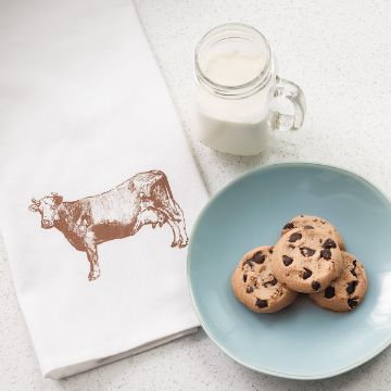 Show details for Etched Cow A jar of milk sits on a white counter next to a light blue plate with three chocolate chip cookies. A white towel with a brown cow illustration is partially visible beside them.