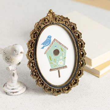 Show details for Birds & Bees An ornate oval frame with a colorful illustration of a green birdhouse, flowers, and a blue bird. A decorative bird figure is beside it on a white surface, with notebooks in the background.