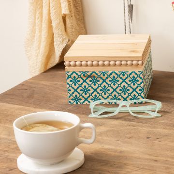 Show details for Moroccan Style A wooden box with an ornate teal pattern sits on a wooden table. Nearby are a pair of light green glasses and a cup of tea on a marble coaster. A cream-colored textured fabric is draped in the background.