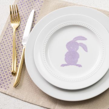Show details for Floral Dot Pattern White plates stacked, top one featuring a purple silhouette of a bunny. Gold fork and knife placed on a beige placemat with a purple pattern. White background.