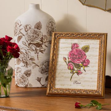 Show details for Music Sheet A wooden table holds a decorative bottle with rose patterns, an ornate framed artwork of roses on sheet music labeled "Rose," a small red rose bouquet in a glass vase, and a single red rose lying on the table.