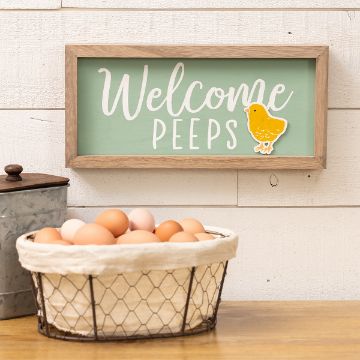 Show details for Welcome Peeps A decorative sign with "Welcome Peeps" and a yellow chick is hung on a white wooden wall. Below it, a wire basket filled with brown eggs sits on a wooden surface.