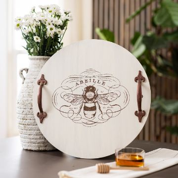 Show details for Honey Bee Round tray with a detailed bee illustration and the word "ABEILLE" in the center, flanked by decorative handles. It rests on a table near a woven vase with white flowers and a small dish of honey, set against a wooden backdrop.