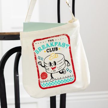 Show details for Breakfast Club A white tote bag hangs on a chair. It features a cartoon of a stack of pancakes with arms and legs, and the text "The Breakfast Club" in colorful letters. The design has a retro style with red, blue, and yellow accents.