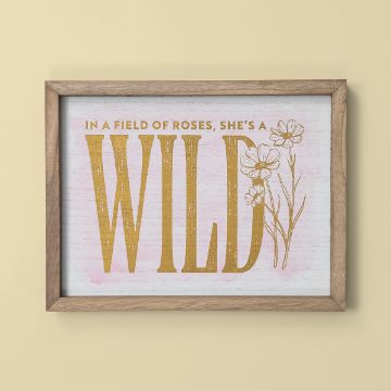 Show details for She's A Wildflower A framed print with the text "In a field of roses, she's a WILD" in gold on a white background. The word "WILD" is large and bold, with line drawings of flowers alongside it. The frame is light wood, and the background is a pale yellow.