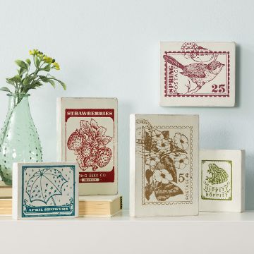 Show details for Spring Postage Decorative art pieces on a shelf display vintage-style prints with botanical, umbrella, and animal designs. A small green vase with foliage is also on the shelf, enhancing the rustic aesthetic against a light blue wall.