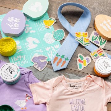 Show details for Egg Hunt Minis A festive Easter crafting setup featuring stencils, stamps, paint jars, and a blue tie decorated with carrots. Various Easter-themed designs include eggs, bunnies, flowers, and a pink onesie reading "Welcome Easter.