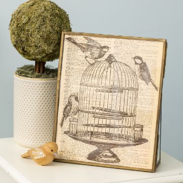 Show details for Birdcage A decorative piece with a vintage illustration of a birdcage and birds on a book page background rests against a light blue wall. Next to it is a small potted, moss-covered topiary and an orange ceramic bird on a white surface.