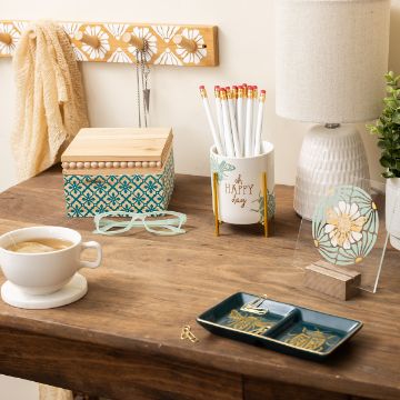 Show details for Oh Happy Day A cozy desk setup with a wooden surface features a ceramic lamp, a patterned box, a mug with pencils, and a cup of tea. A pair of glasses, a decorative plate, and a small dish with trinkets add to the ambiance. A scarf hangs from a peg rack.