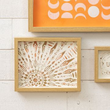 Show details for Sunburst Pattern Framed artwork on a white wall, with a wooden frame. The art features a radial pattern in brown and gold tones, with geometric shapes and intricate designs. Orange artwork partially visible above.