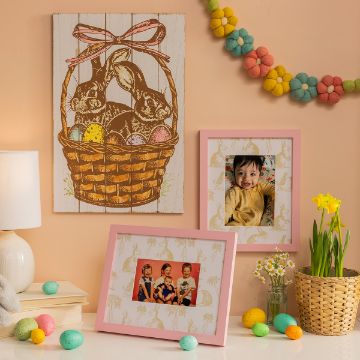 Show details for Bunny Basket A cozy Easter-themed scene with framed family photos, a rabbit and egg wall art, colorful decorative eggs, a table lamp, and a pot of daffodils. Vibrant felt flowers hang above, creating a cheerful holiday atmosphere.