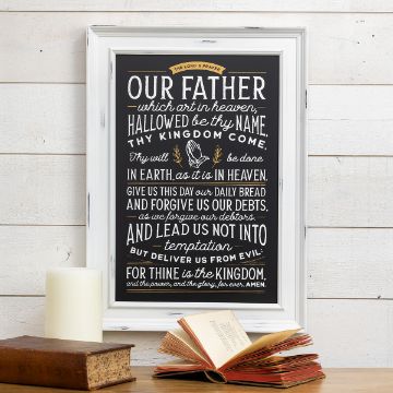 Show details for The Lord's Prayer A framed chalkboard-style print displaying "The Lord's Prayer" in stylish lettering. Below it, a closed book and a lit candle are placed on a wooden surface. The background is a white wooden wall.