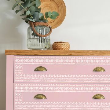 Show details for Watercolor Pattern A pink dresser with brass handles features intricate white geometric patterns. A wooden plate, small woven basket, and a clear vase with green eucalyptus branches rest on the wooden top. The background wall is light-colored.