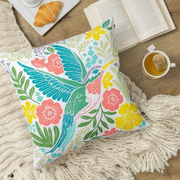 Show details for Hummingbird A colorful pillow with a bird and floral design is on a cozy knitted blanket on a wooden floor. Nearby are an open book, a plate with a croissant, and a cup of tea.