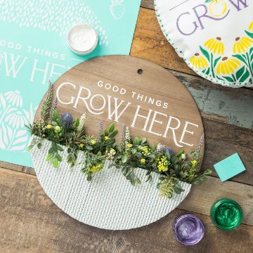 Show details for Good Things Grow Here A round wooden sign with "Good Things Grow Here" text, adorned with artificial green foliage and yellow flowers, lies on a wooden surface. Nearby are green and purple paints, decorative papers, and a pillow with a floral design.