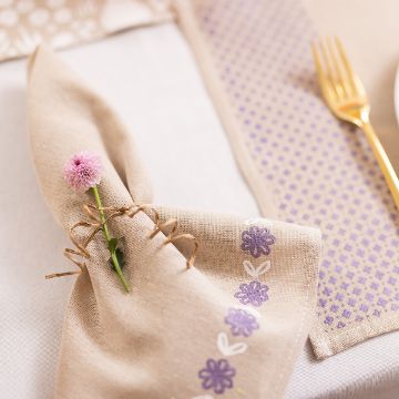 Show details for Natural Napkin (2-Pack, Tan, 18" x 18") A beige napkin adorned with purple floral designs is wrapped around a pink flower and tied with twine. It rests on a table next to a golden fork and a patterned lavender table runner.